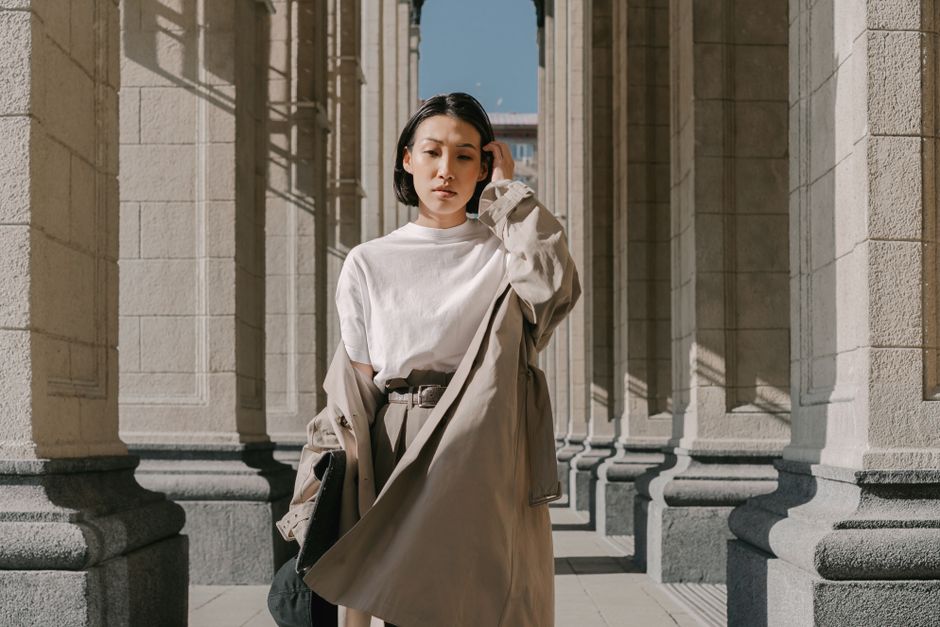 Stylish Asian woman wearing a trench coat stands confidently in an elegant architectural setting.