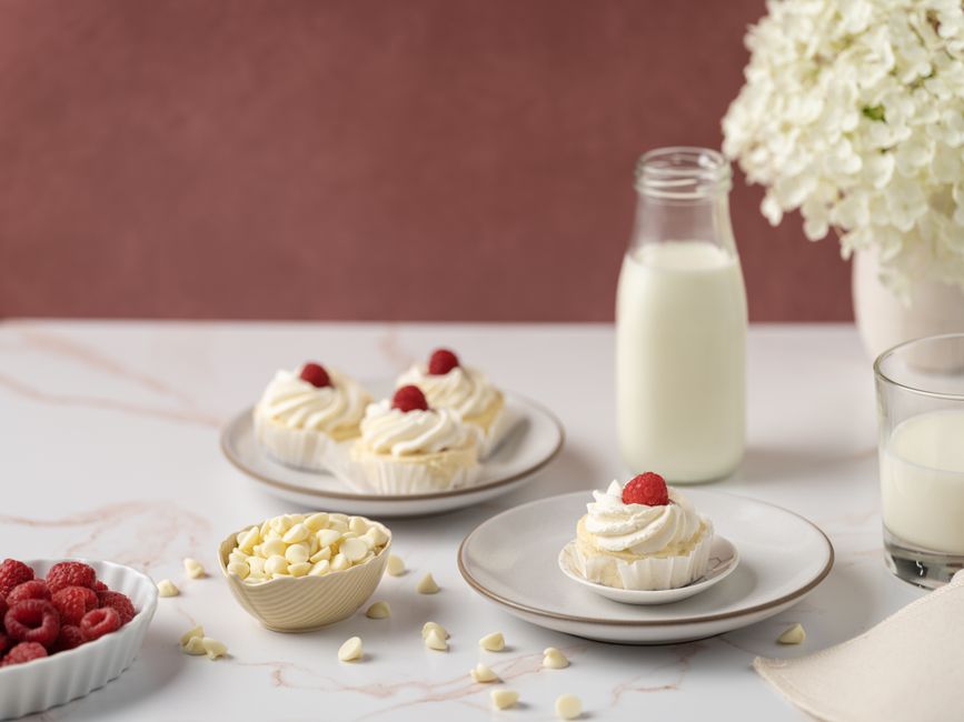 A lovely display of vanilla cupcakes topped with raspberries, accompanied by milk and white chocolate chips.
