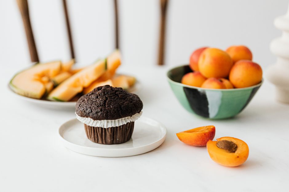 High angle of yummy chocolate cupcake in white ceramic plate placed on table with bowl of ripe apricots and sliced melons