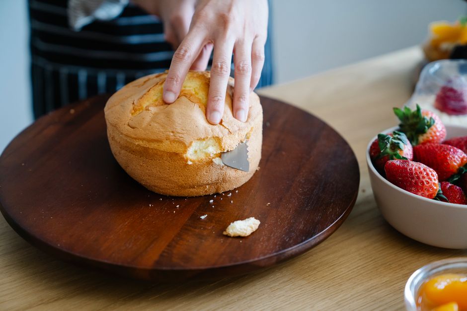 Hand slicing a sponge cake on a wooden platter with fresh strawberries nearby.