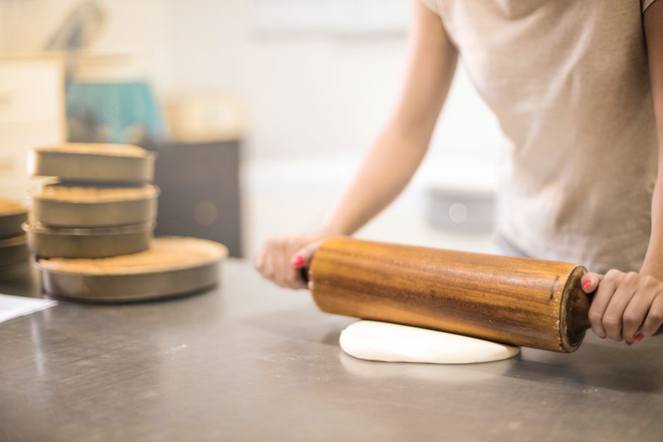 A woman using a wooden rolling pin to flatten dough in a bakery kitchen setting.