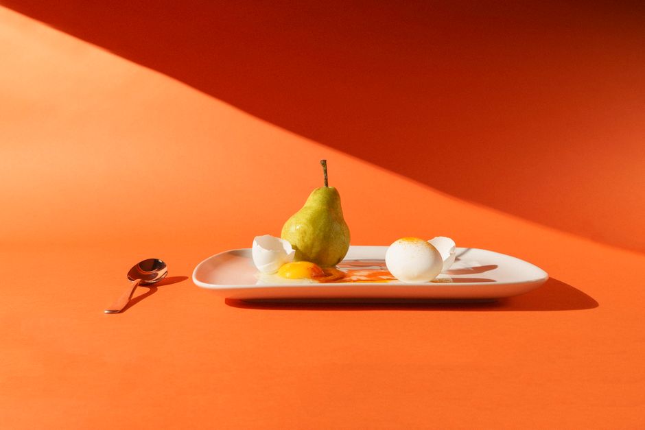 Artistic still life featuring a pear and cracked eggs on a plate against a vivid orange background.