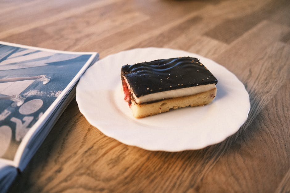 A slice of chocolate and cream cake on a white plate, next to a magazine on a wooden table.