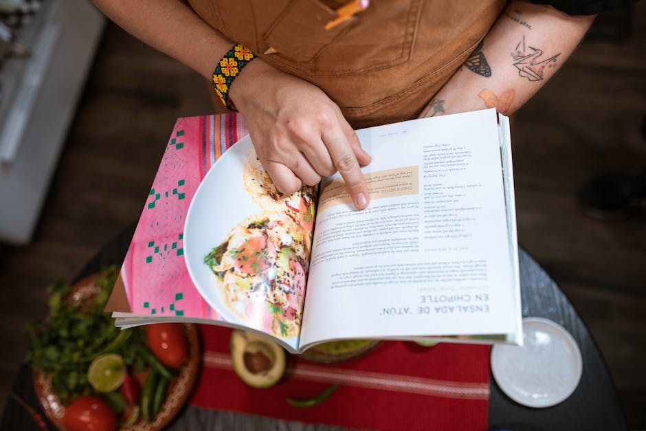 Close-up of a person pointing at a recipe in an open cookbook next to fresh ingredients.